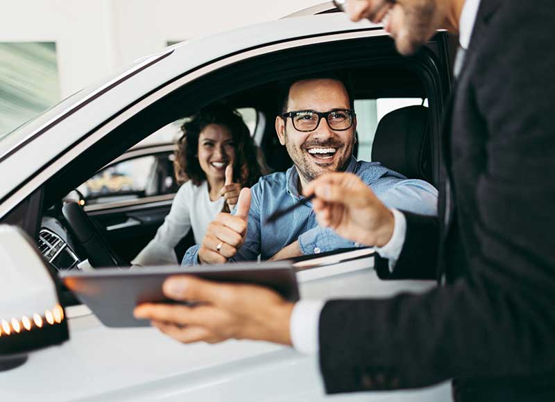 Smiling car buyers sitting in a vehicle giving a thumbs-up while a salesperson shows them details on a tablet.